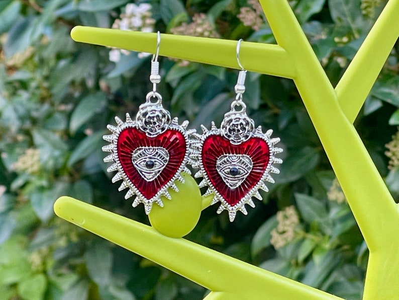 May include: A pair of silver earrings with red heart-shaped charms. Each charm features a silver eye with a black pupil and a silver floral design at the top.