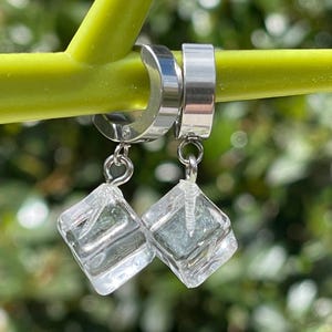 May include: A pair of silver-toned metal hoop earrings, each featuring a dangling clear, cube-shaped charm that resembles an ice cube. The earrings are suspended from a smooth, polished metal hoop mechanism with a secure hinged closure.