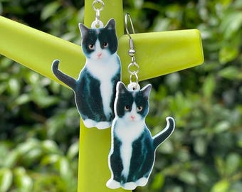 May include: A pair of drop earrings featuring a realistic graphic print of a black and white tuxedo cat. Each earring is suspended from a silver-toned metal hook and displays the cat in a seated position with a white chest and paws.