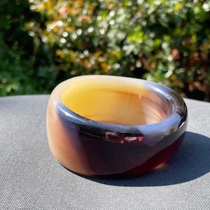May include: A chunky, polished ring with a rounded shape. The ring features a gradient of colors, including yellow, brown, and deep purple. The ring is sitting on a gray surface, with a blurred green background.
