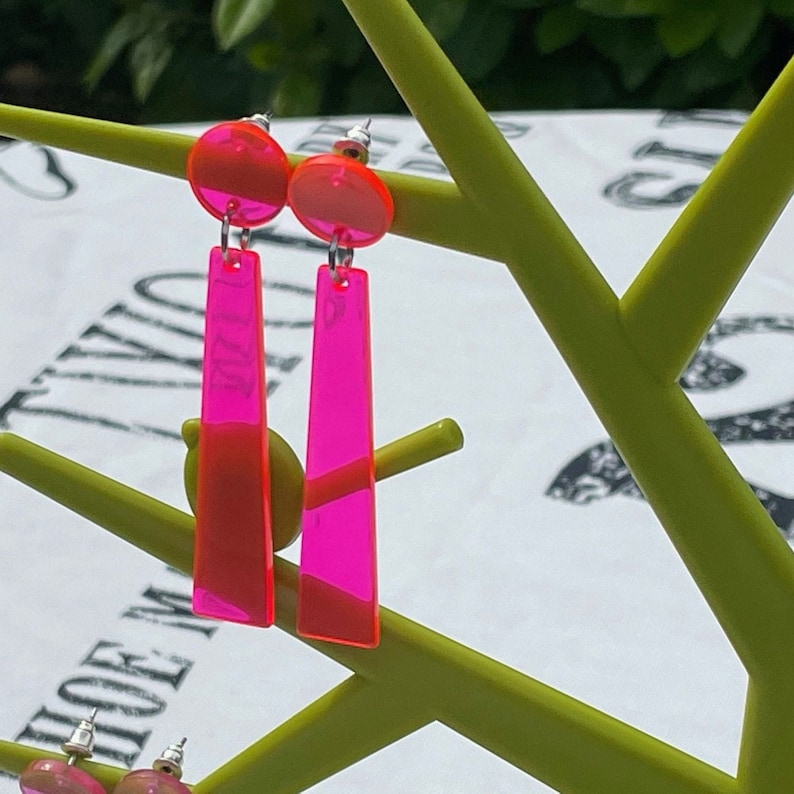 May include: A pair of pink acrylic earrings with a long, rectangular shape. The earrings are hanging from a green display stand.