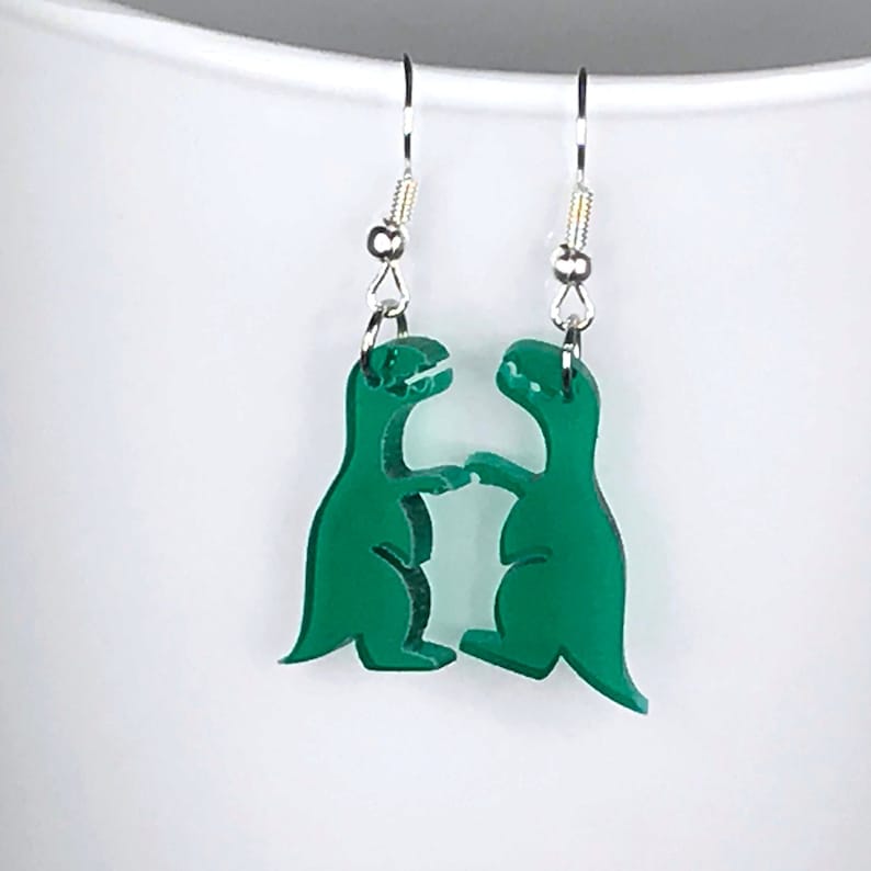 May include: A pair of green acrylic earrings shaped like two dinosaurs facing each other. The dinosaurs are in a fighting pose with their arms raised.