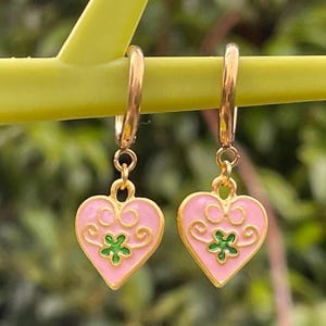 May include: Gold-coloured hoop earrings with pink heart-shaped pendants. Each pendant has a green floral design and gold-coloured filigree. The earrings are shown against a blurred green background.