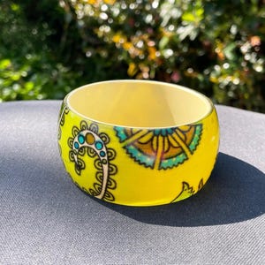 May include: A yellow bangle bracelet featuring a floral paisley pattern in teal, blue, and black. The bracelet, made of a glossy material, rests on a grey surface. The inside of the bracelet is a lighter yellow.