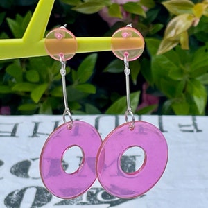 May include: A pair of pink and clear acrylic earrings with a donut shape. The earrings have a silver hook and a small clear circle at the top.
