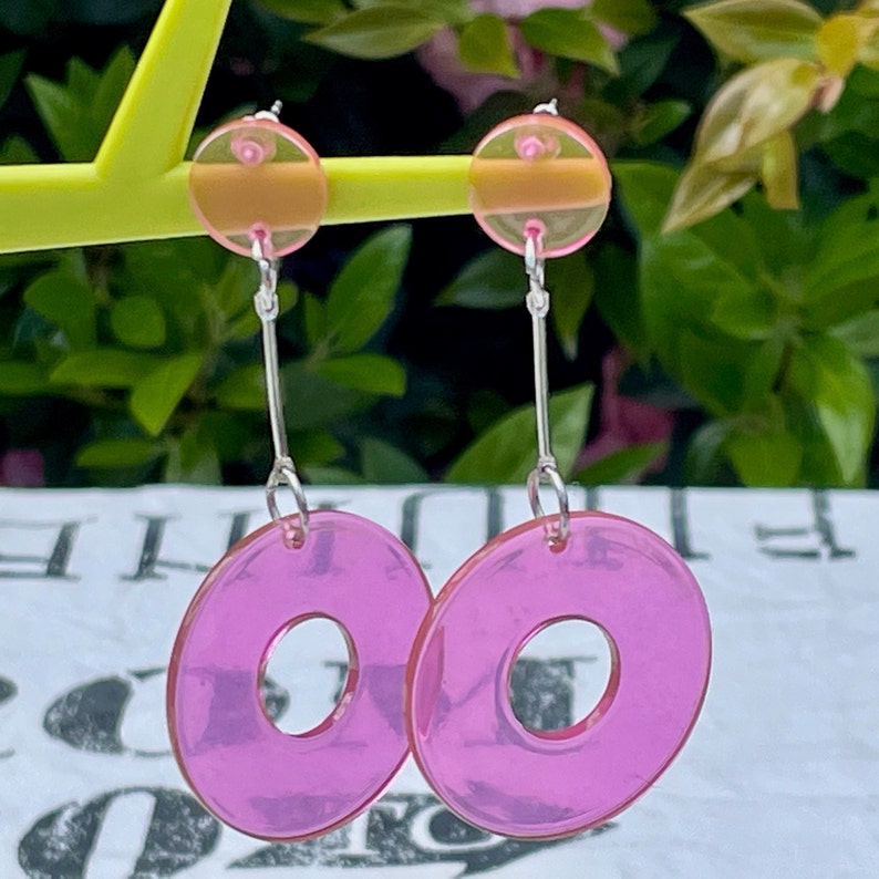 May include: A pair of pink and clear acrylic earrings with a donut shape. The earrings have a silver hook and a small clear circle at the top.