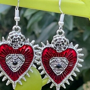 May include: A pair of silver earrings with red heart-shaped charms. Each charm features a silver eye with a black pupil and a silver rose design at the top.