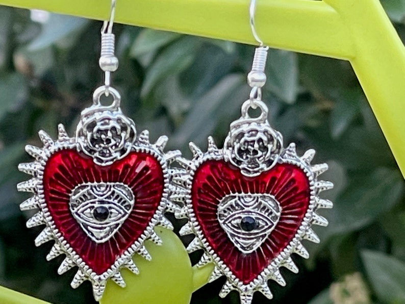 May include: A pair of silver earrings with red heart-shaped charms. Each charm features a silver eye with a black pupil and a silver rose design at the top.