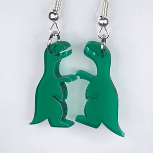 May include: A pair of green dinosaur earrings. The dinosaurs are facing each other and appear to be shaking hands. The earrings are made of acrylic and have silver hooks.