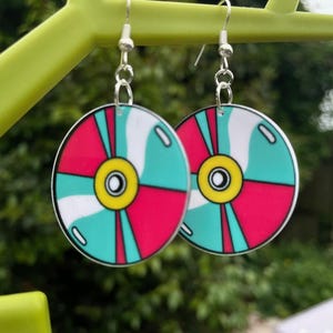 Può includere: Un paio di orecchini rotondi con un design colorato. Ogni orecchino presenta un motivo turchese, rosa e bianco con un centro giallo. Gli orecchini sono sospesi a ganci color argento.