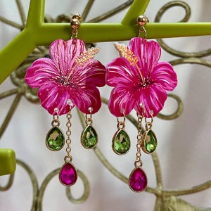 Może przedstawiać: Kolczyki w kształcie różowego kwiatu hibiskusa z zielonymi i różowymi akcentami kryształowymi. Kolczyki są wiszące i mają złoty łańcuszek.