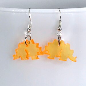 May include: A pair of orange dinosaur earrings with silver hooks. The dinosaurs are facing each other and are made of a translucent material.