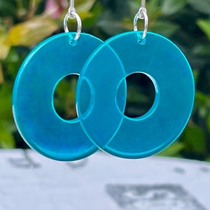 May include: A pair of turquoise blue acrylic earrings with a donut shape. The earrings have a silver hook.