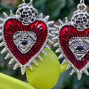 May include: A pair of silver earrings with red enamel hearts. Each heart has a silver eye with a black pupil in the center. The hearts are surrounded by a silver halo with spikes.