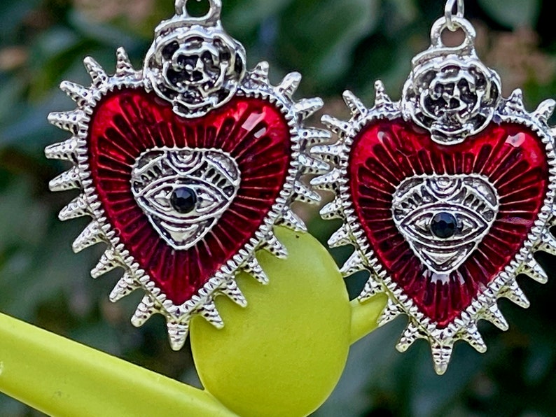 May include: A pair of silver earrings with red enamel hearts. Each heart has a silver eye with a black pupil in the center. The hearts are surrounded by a silver halo with spikes.