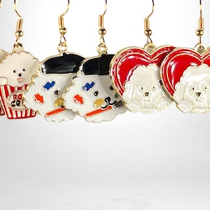 May include: Six pairs of gold dangle earrings with different charms. The charms include a white dog with a red and white striped popcorn bucket, a white dog with a black beret and paintbrush, and a white dog inside a red heart.