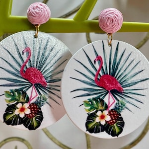 Może przedstawiać: Para okrągłych drewnianych kolczyków w kolorze różowym i białym z motywem flaminga i tropikalnych liści. Kolczyki są okrągłe i mają różową kwiatową ozdobę z tkaniny na górze.