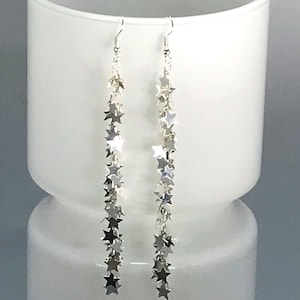 May include: Silver dangle earrings featuring multiple star charms. The earrings have a hook closure and the stars are strung together in a cascading design. The earrings are displayed against a white background.
