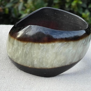 May include: A handcrafted, oval-shaped bracelet in shades of black, brown, and white. The bracelet has a glossy finish and a unique marbled pattern. The design is a blend of modern and organic aesthetics.