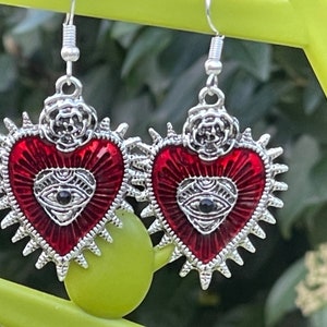 May include: A pair of silver earrings with red enamel hearts. Each heart has a silver eye with a black pupil and a silver rose on top.