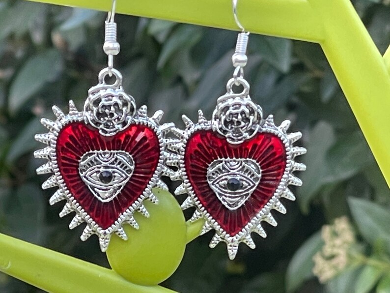 May include: A pair of silver earrings with red enamel hearts. Each heart has a silver eye with a black pupil and a silver rose on top.