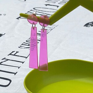 May include: A pair of pink acrylic earrings with a long, rectangular shape. The earrings are hanging from a green display stand.