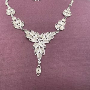 May include: A silver filigree necklace with a floral pendant and leaf-shaped accents. The necklace features intricate openwork designs and a delicate chain. The jewelry is displayed on a purple surface.