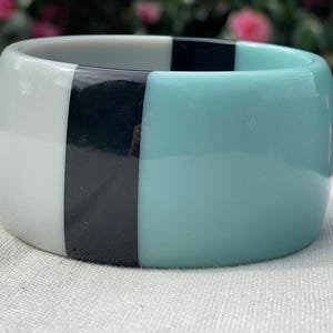 May include: A wide, smooth bangle bracelet featuring a colour-blocked design with three distinct vertical sections in solid white, glossy black, and soft light blue. The bracelet has a thick, rounded profile and a polished, reflective finish.