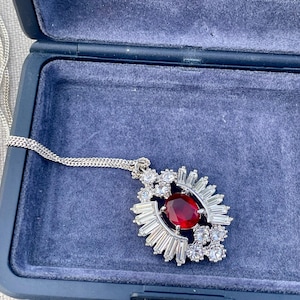 May include: A silver necklace with a large, oval, red gemstone pendant. The pendant is surrounded by smaller, clear gemstones and baguette-cut stones. The necklace is displayed in a blue jewelry box.