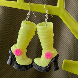 May include: A pair of neon yellow boot earrings with black heels and pink rose accents. The earrings are suspended from silver hooks and feature a ridged design on the boot shaft. The heels have a platform sole.