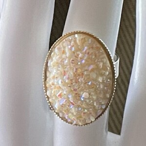 May include: An oval-shaped ring featuring a textured, cream-colored druzy stone with iridescent, shimmering highlights. The stone is set within a thin, metallic gold-toned bezel frame that follows the oval contour of the central gemstone piece.
