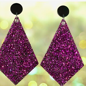 May include: A pair of dangle earrings featuring large, diamond-shaped pendants covered in vibrant purple glitter. Each pendant is attached to a small, solid black circular stud component by a silver-toned metal jump ring, creating a simple drop design.