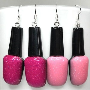 Può includere: Quattro paia di orecchini pendenti a forma di bottiglie di smalto per unghie. Due bottiglie sono rosa con glitter e due bottiglie sono di un magenta intenso con glitter.
