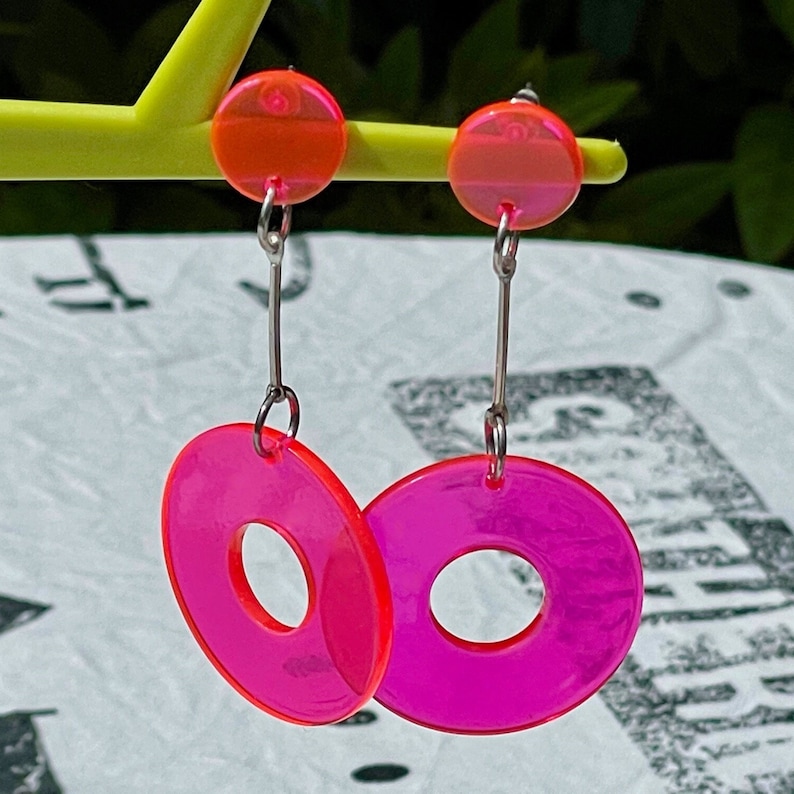 May include: A pair of pink and orange dangle earrings with a silver chain and a round, hollow pink disc.