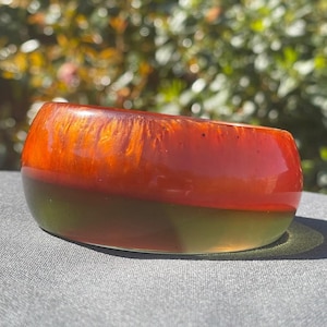 May include: A handcrafted resin bowl with a two-tone design. The top half is a vibrant orange, transitioning to a deep olive green at the base. The bowl has a smooth, glossy finish and is ideal for decorative use.