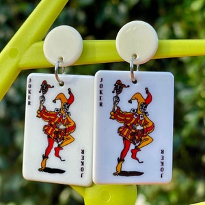 Può includere: Un paio di orecchini bianchi a forma di carta con un'immagine di un joker su ciascuno. Il joker indossa un costume da giullare rosso, giallo e verde e tiene un bastone con una testa di giullare su di esso. Il testo "JOKER" è stampato su ogni carta.