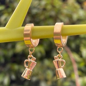 May include: Gold-coloured hoop earrings with small coffee pot charms. The earrings are suspended from a green stem, with a blurred green background. The coffee pot charms have a handle and spout.