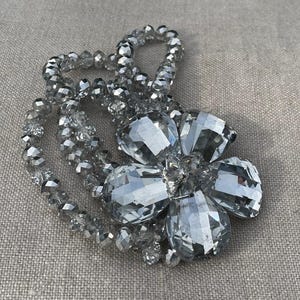 May include: A silver-toned necklace featuring a large, faceted flower pendant. The pendant is surrounded by a beaded strand of smaller, similarly faceted beads. The necklace is displayed on a neutral, textured surface.