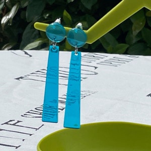 May include: A pair of turquoise blue dangle earrings with a round turquoise blue top. The earrings are hanging from a green surface.