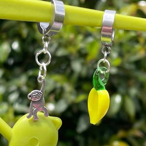 May include: A pair of silver hoop earrings with dangling charms. One earring features a small, silver monkey charm. The other earring has a yellow banana charm with a green stem. The earrings are displayed against a blurred green background.
