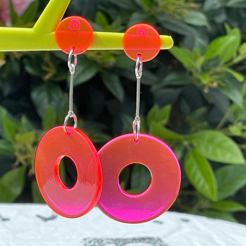 May include: A pair of pink and red hoop earrings with a silver chain. The earrings are hanging from a yellow stand.