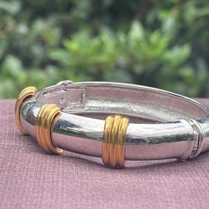 Könnte beinhalten: Silberfarbenes Armband mit goldenen Akzenten. Das Armband hat eine polierte Silberoberfläche mit drei goldenen Bändern. Es liegt auf einer mauvefarbenen Oberfläche.