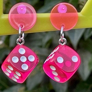 May include: A pair of pink dice earrings with white dots. The dice are dangling from silver hooks attached to pink circular charms.