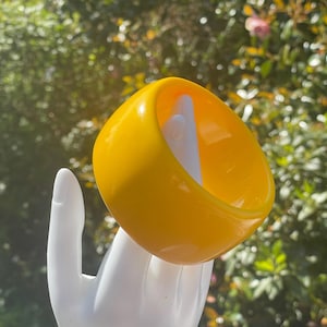 May include: A vibrant yellow, square bangle bracelet with a white interior. The bracelet is held up by a white hand, with a blurred green background of foliage.