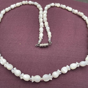 May include: A white beaded necklace with a magnetic clasp. The necklace features a repeating pattern of small, flower-shaped beads interspersed with smaller, round beads. The necklace is displayed on a purple background.