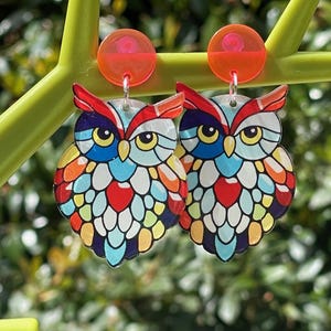 May include: A pair of owl-shaped earrings with a stained-glass effect. The earrings are in various colours, including red, blue, and yellow. Each earring has a round, orange top. The earrings are suspended against a blurred green backdrop.