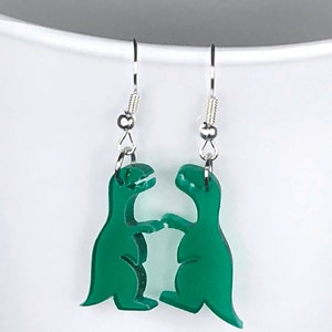 May include: A pair of green dinosaur earrings with silver hooks. The dinosaurs are facing each other and appear to be dancing.