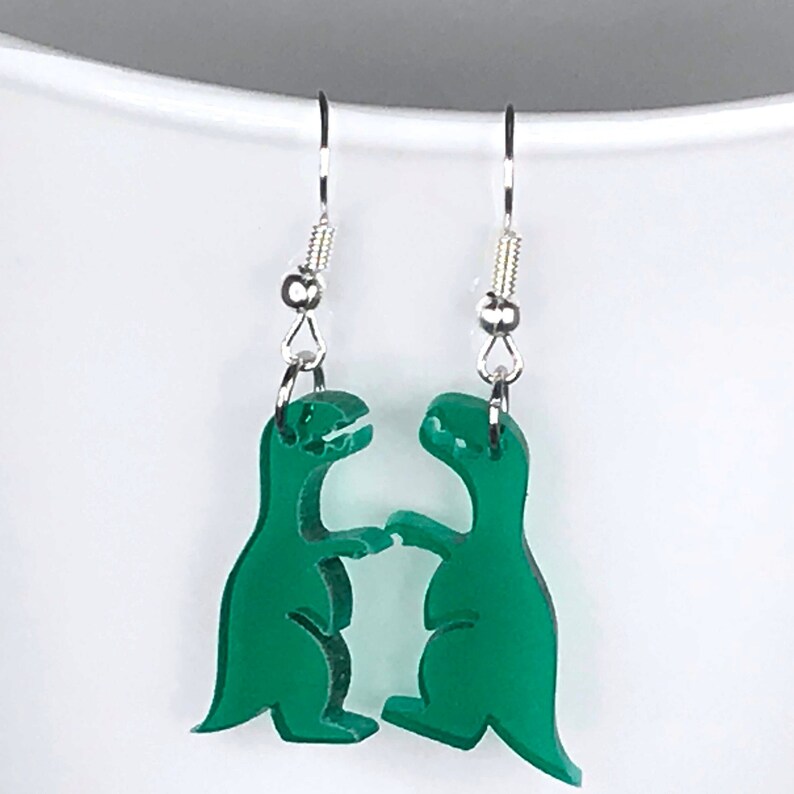 May include: A pair of green dinosaur earrings with silver hooks. The dinosaurs are facing each other and appear to be dancing.
