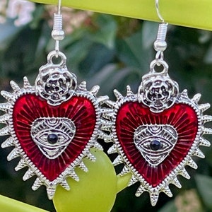 May include: A pair of silver earrings with red heart-shaped charms. Each charm features a silver eye with a black pupil and a silver rose on top. The hearts are surrounded by silver spikes.
