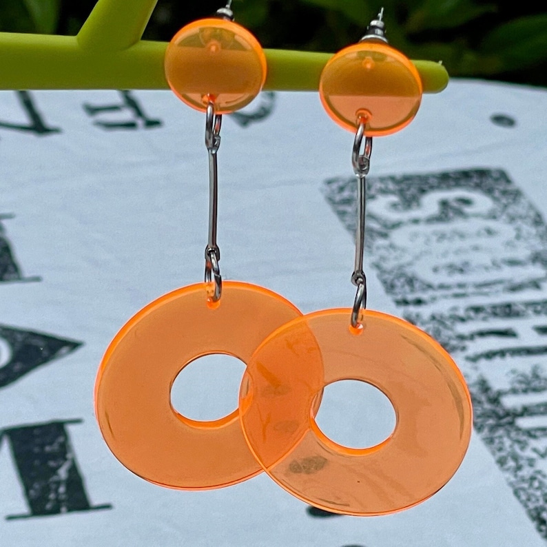May include: A pair of orange acrylic earrings with a donut shape. The earrings are suspended from silver chains.